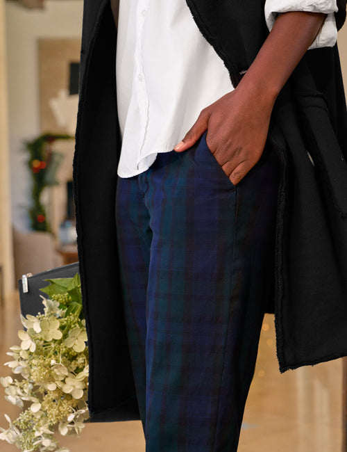 A person wearing dark blue and green plaid trousers, a white button-up shirt, and a black jacket, standing indoors with one hand in their pocket., view 3