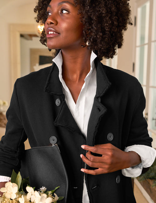 A woman wearing a black double-breasted blazer over a white collared shirt, holding a black clutch and white flowers in an indoor setting., alternate view