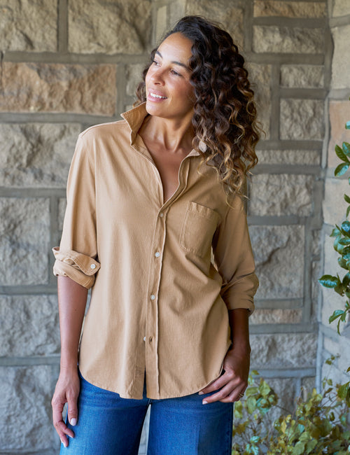 A woman wearing a tan long-sleeve button-up shirt with rolled sleeves and blue jeans, standing in front of a stone wall., front view