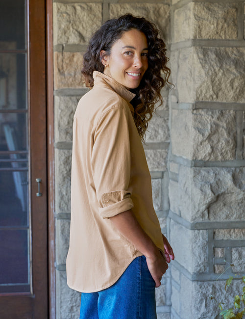 A woman with curly dark hair stands on a stone porch wearing a beige button-up shirt with rolled sleeves and blue jeans, looking back over her shoulder., alternate view