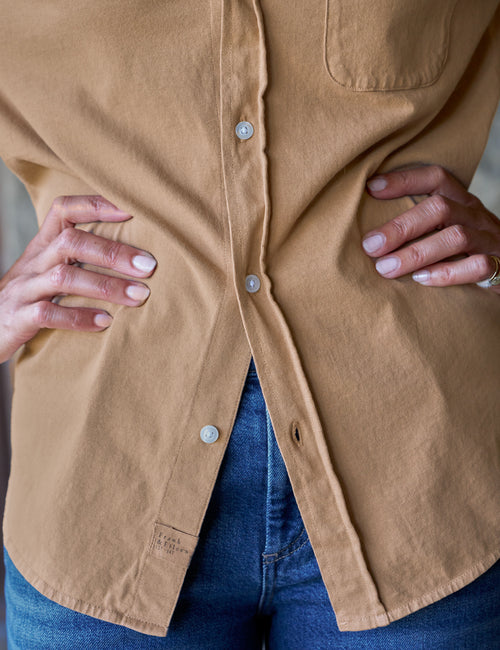 A close-up of a person wearing a tan button-front overshirt with a chest pocket and blue jeans, with both hands resting on the waist., view 3