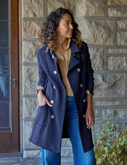 A woman stands on a stone patio wearing a navy blue double-breasted coat over a tan top and wide-leg blue jeans, with a stone wall and potted plants in the background., front view