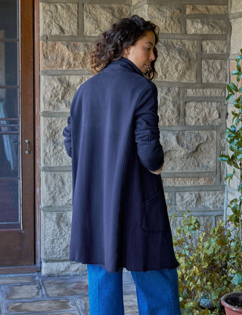 A person standing on a stone patio in front of a stone wall, wearing a dark navy long coat over blue wide-leg jeans and brown shoes, with a potted plant to the right., alternate view