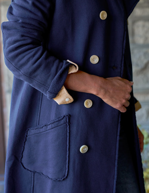 A close-up of a person wearing a navy blue coat with large cream buttons and a visible tan lining at the cuff, shown against a softly blurred stone background., view 3