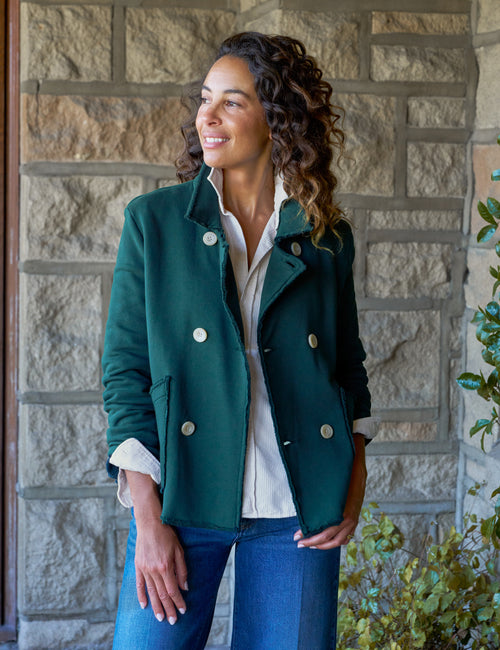 A woman wearing a dark green double-breasted jacket over a white shirt and blue jeans, standing in front of a stone wall., front view