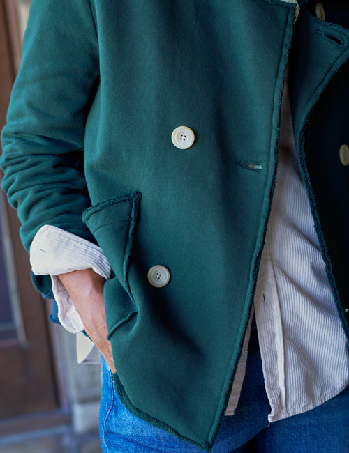 A close-up of a person wearing a dark green double-breasted jacket , layered over a white shirt and blue jeans., view 3