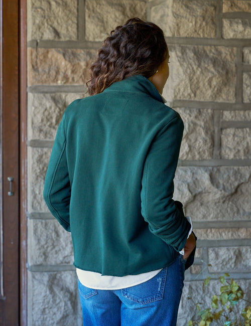 A person viewed from behind wearing a dark green fleece jacket over a white shirt and blue jeans, standing in front of a stone wall., alternate view