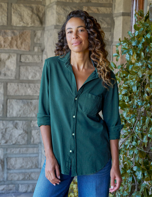 A woman with curly brown hair wearing a dark green button-down shirt and blue jeans, standing in front of a stone wall and green ivy., front view