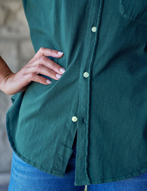 Close-up of a person wearing a dark teal-green button-up shirt with white buttons and blue jeans, with one hand resting on the hip., view 3