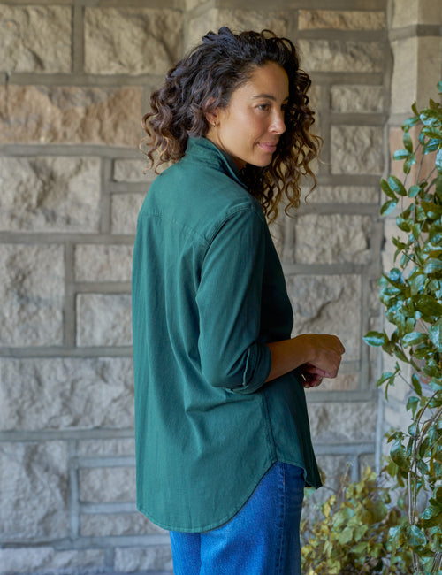 A woman with curly brown hair is shown from the back and side, wearing a dark green long-sleeve button-up shirt and blue jeans, standing in front of a stone wall with green plants on the right., alternate view