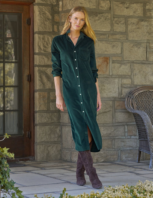 A woman stands on a stone patio wearing a dark green corduroy button-front maxi dress with long sleeves and brown suede boots. A stone wall, a glass door, and a wicker chair are visible in the background., front view