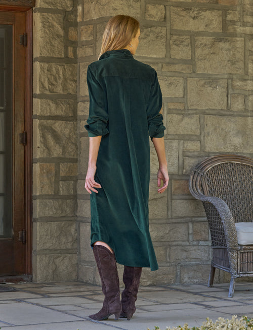 A woman stands with her back to the camera wearing a dark green corduroy maxi shirt dress and brown knee-high boots, posing in front of a stone wall with a wicker chair to the side., alternate view