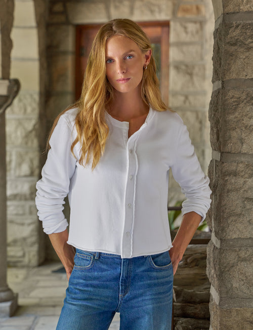 A woman with long blonde hair wearing a white long-sleeve button-up shirt and blue jeans, standing in front of a stone column., front view