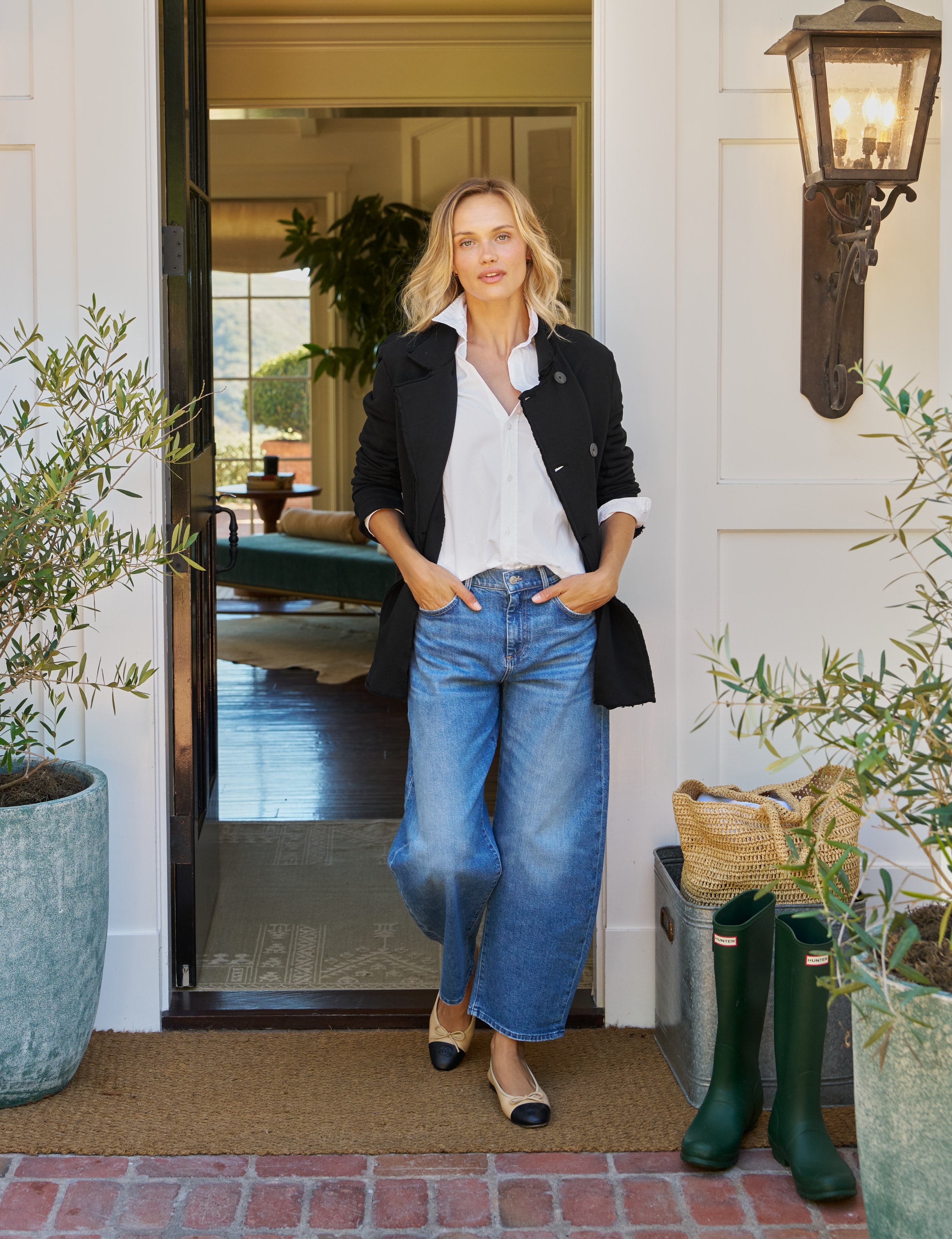 Woman standing outside a house with plants and green boots.