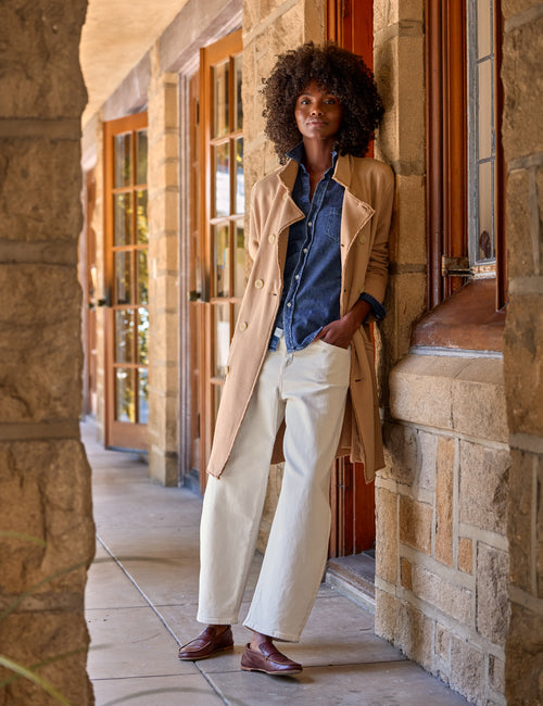 A woman stands on a stone walkway wearing a beige trench coat over a blue denim shirt and white wide-leg trousers, with brown leather loafers., view 5