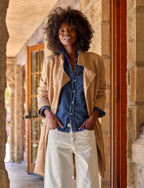 A woman stands on a stone walkway wearing a beige trench coat over a blue button-up shirt, cream wide-leg pants, and dark brown loafers., front view