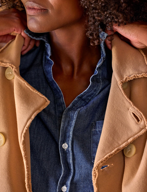 Close-up of a person wearing a tan  coat with raw, frayed edges over a blue denim button-up shirt. The person's lower face, neck, and hands are visible., view 3