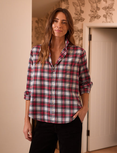 A woman wearing a red, white, and black plaid button-down shirt with rolled-up sleeves, paired with black pants, standing in an indoor setting with patterned wallpaper and a white door in the background., front view