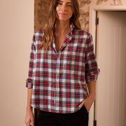 A woman wearing a red, white, and black plaid button-down shirt with rolled-up sleeves, paired with black pants, standing in an indoor setting with patterned wallpaper and a white door in the background.