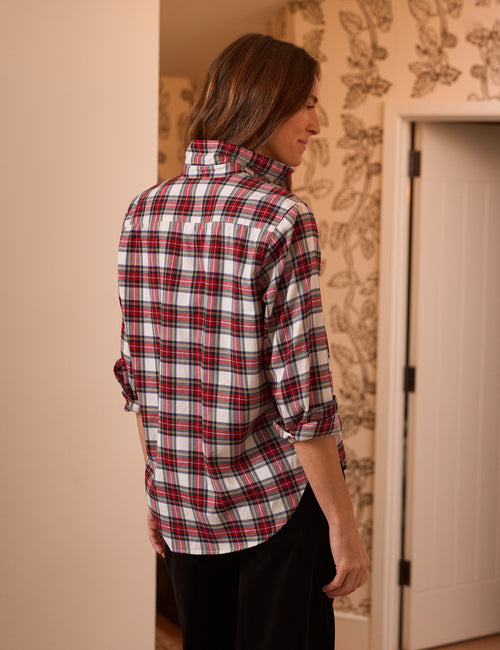 A woman shown from the back wearing a red, white, and black plaid button-up shirt with rolled-up sleeves, standing in an indoor setting with patterned wallpaper., alternate view