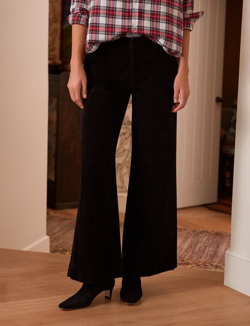 A person wearing black wide-leg pants with a red, white, and black plaid button-up shirt and black pointed-toe heeled boots, standing in a home interior with wood floors., front view