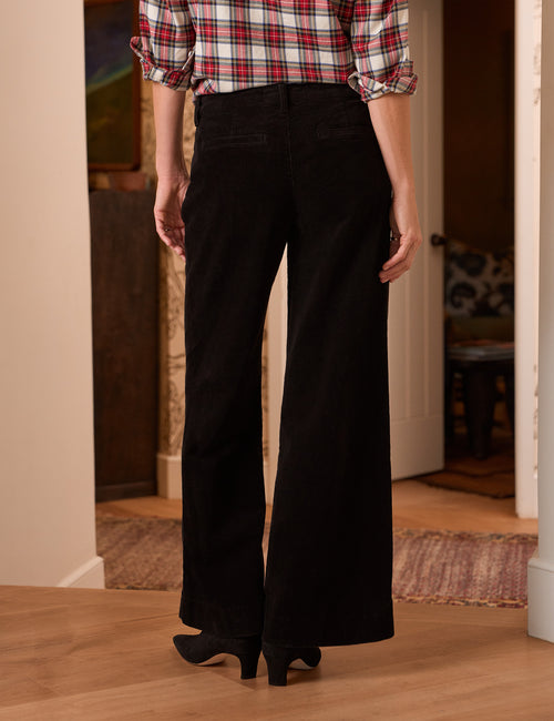 A person shown from the back wearing black wide-leg pants, a red, white, and black plaid shirt, and black heels in an indoor room with wood floors., alternate view