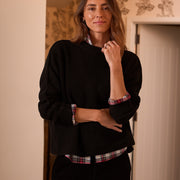 A woman wearing a black ribbed knit sweater over a red, white, and green plaid button-down shirt, paired with black pants, standing in a warmly lit indoor setting.