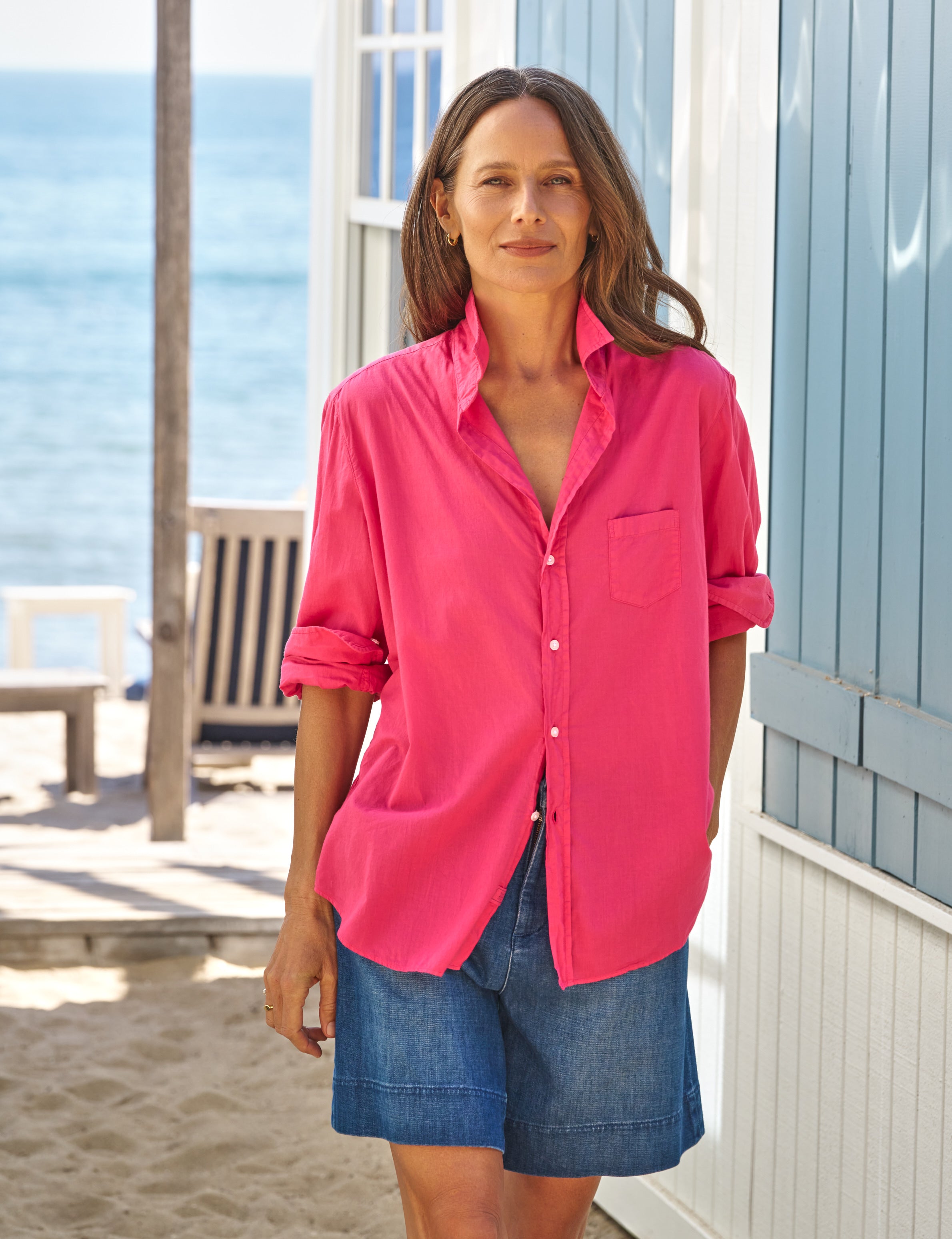 Woman wearing a bright pink shirt and blue shorts standing on a beach., front view