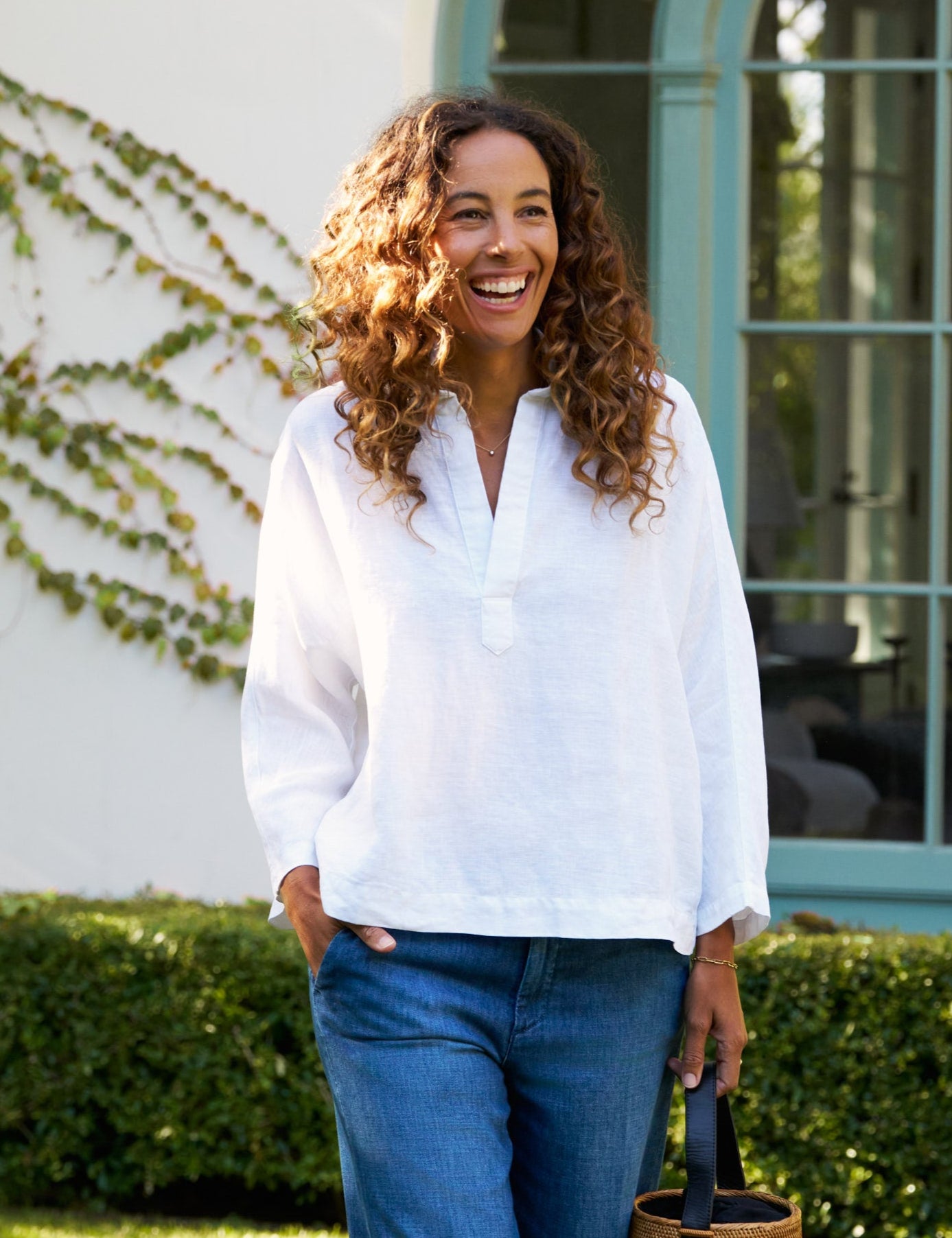 Woman in white shirt and blue jeans walking outdoors with a basket, smiling.