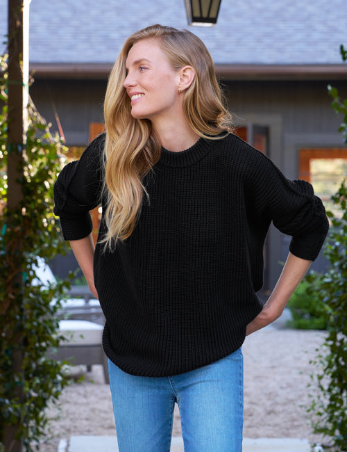 A woman wearing a black Montecito Sweater paired with blue jeans, standing outdoors., front view