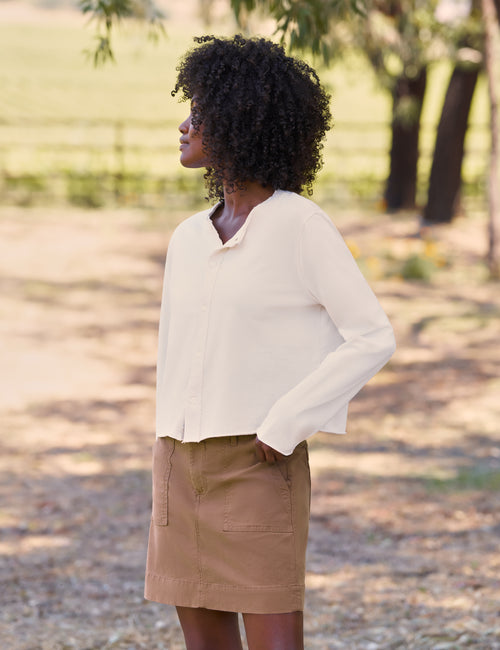A woman wearing a cream long-sleeve button-up blouse and a brown mini skirt, standing outdoors in a sunny, blurred park-like setting., view 4