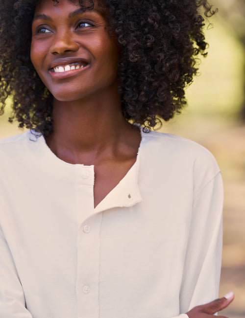 A smiling woman with curly dark hair wearing a light cream-colored long-sleeve button-up shirt, photographed outdoors against a softly blurred green and beige background., view 3