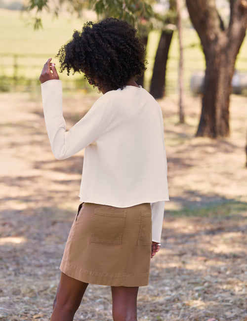 A woman photographed from behind outdoors, wearing a cream long-sleeve cropped top and a tan mini skirt in a park-like setting with trees and dry ground., alternate view