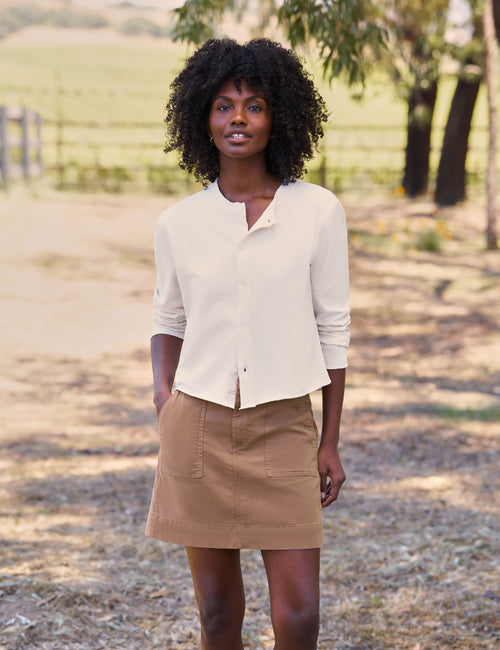 A woman standing outdoors wearing a cream-colored, cropped long-sleeve button-up shirt and a tan mini skirt., view 5