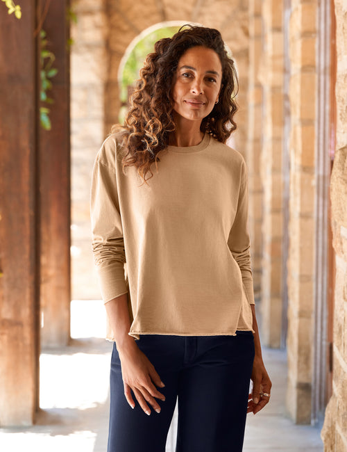 A woman with curly brown hair wearing a beige long-sleeve crewneck top and dark navy pants, standing in a stone archway corridor., front view