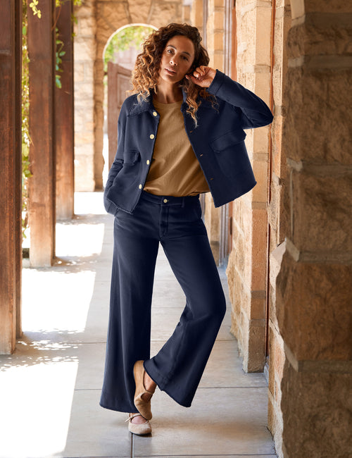 A woman stands in a sunlit stone corridor wearing a navy blue cropped jacket and matching wide-leg pants over a tan top, with tan flats., view 5
