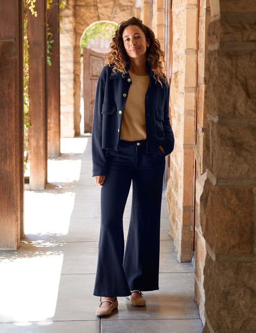 A woman stands in a sunlit stone walkway wearing a navy blue jacket and matching wide-leg pants over a tan top, with tan shoes., front view