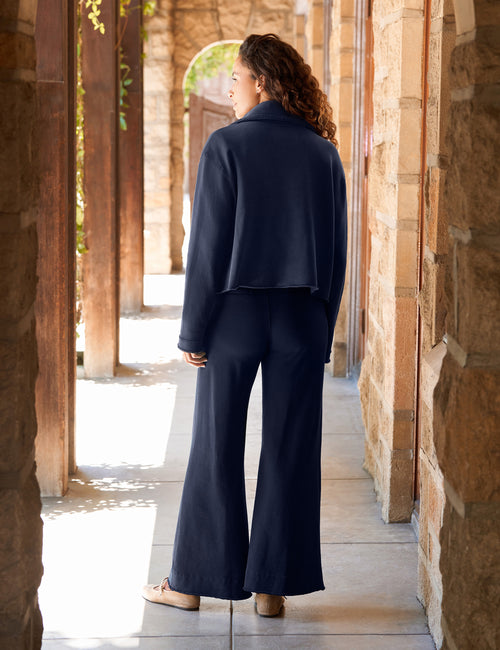 A woman stands in a sunlit stone corridor wearing a navy blue cropped jacket and matching wide-leg pants, shown from the back., alternate view