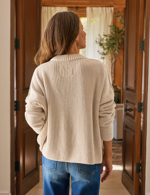 Woman wearing a beige sweater and blue jeans standing in a doorway., alternate view