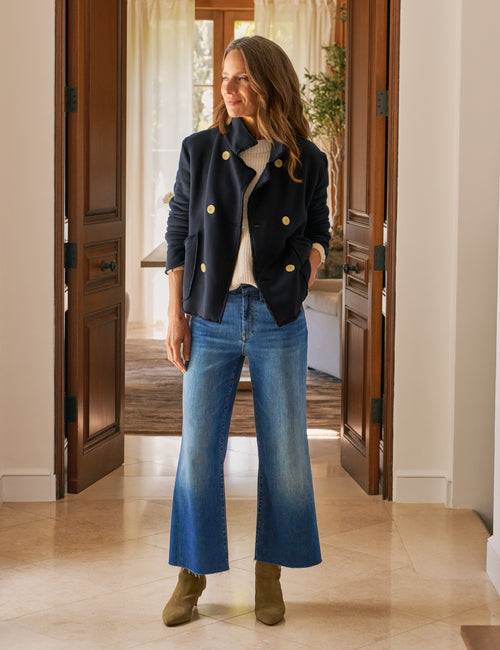 Woman wearing a navy jacket with gold buttons, white sweater, blue jeans, and green shoes in a home setting., view 5