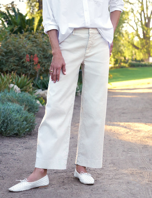 Person wearing a white shirt and light-colored pants standing outdoors with greenery in the background, front view