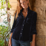 A woman with long light brown hair leans against a rustic stone pillar in an outdoor patio setting, wearing a dark navy blue velvet button-down shirt and blue jeans.