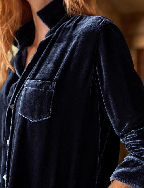 Close-up of a person wearing a dark navy velvet long-sleeve button-up shirt with a chest pocket, shown against a softly blurred warm-toned indoor background., view 3