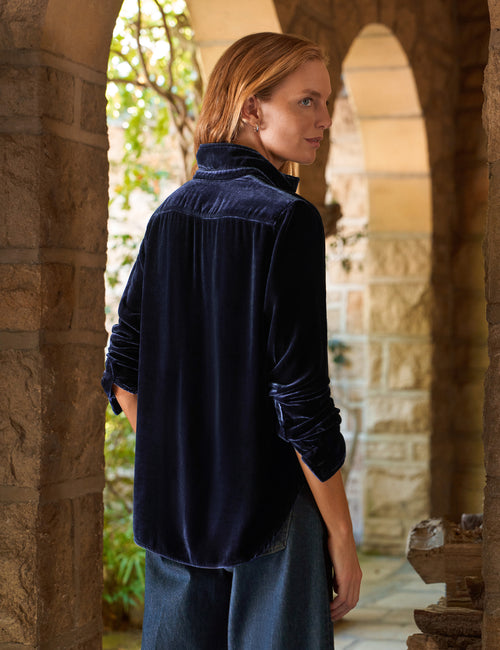 A woman wearing a dark navy blue velvet long-sleeve shirt and wide-leg blue jeans, standing in a stone archway corridor., alternate view