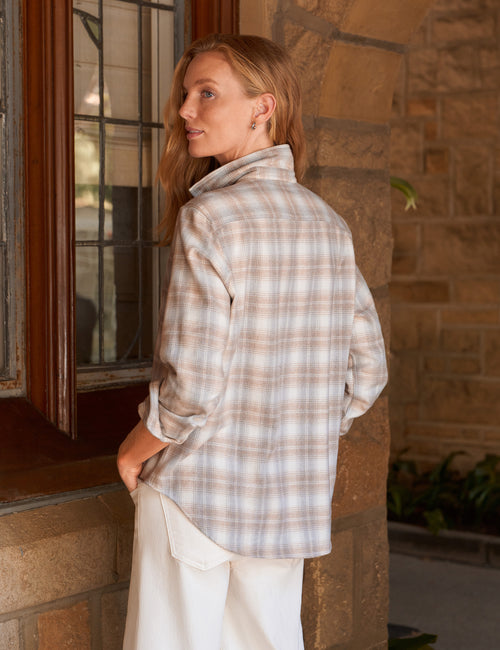 A woman wearing a beige and white plaid button-down shirt and white pants, standing in profile next to a rustic stone wall and window., alternate view