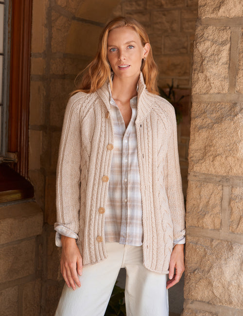 A woman stands in a stone doorway wearing a cream cable-knit button-front cardigan over a light plaid button-up shirt and white pants., front view