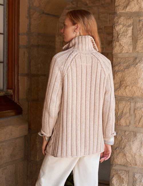 A woman wearing a cream ribbed turtleneck sweater and white pants, photographed from the back in a stone-walled interior setting., alternate view