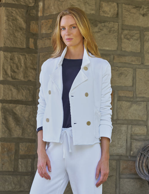 A woman stands outdoors against a stone wall wearing a white double-breasted jacket with gold-tone buttons, a dark navy top, and white drawstring pants., front view