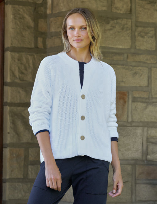 A woman wearing a white ribbed knit cardigan, paired with dark pants, standing in front of a stone wall., front view