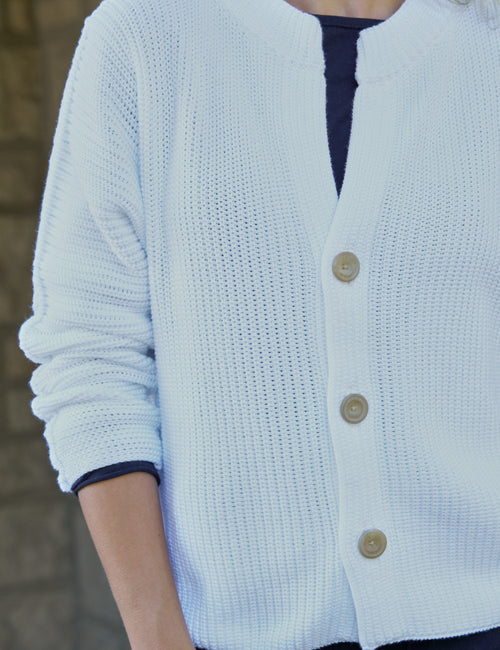 A close-up of a person wearing a white cardigan with three buttons over a dark navy top. The background is a softly blurred stone wall., view 3